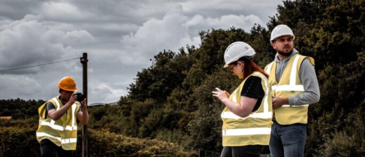 Sam Brown taking a photo of Amy Corker and Lewis Jarvis operating a DJI drone camera on a construction site. Everyone is wearing white safety hard hats and high visibility jackets.