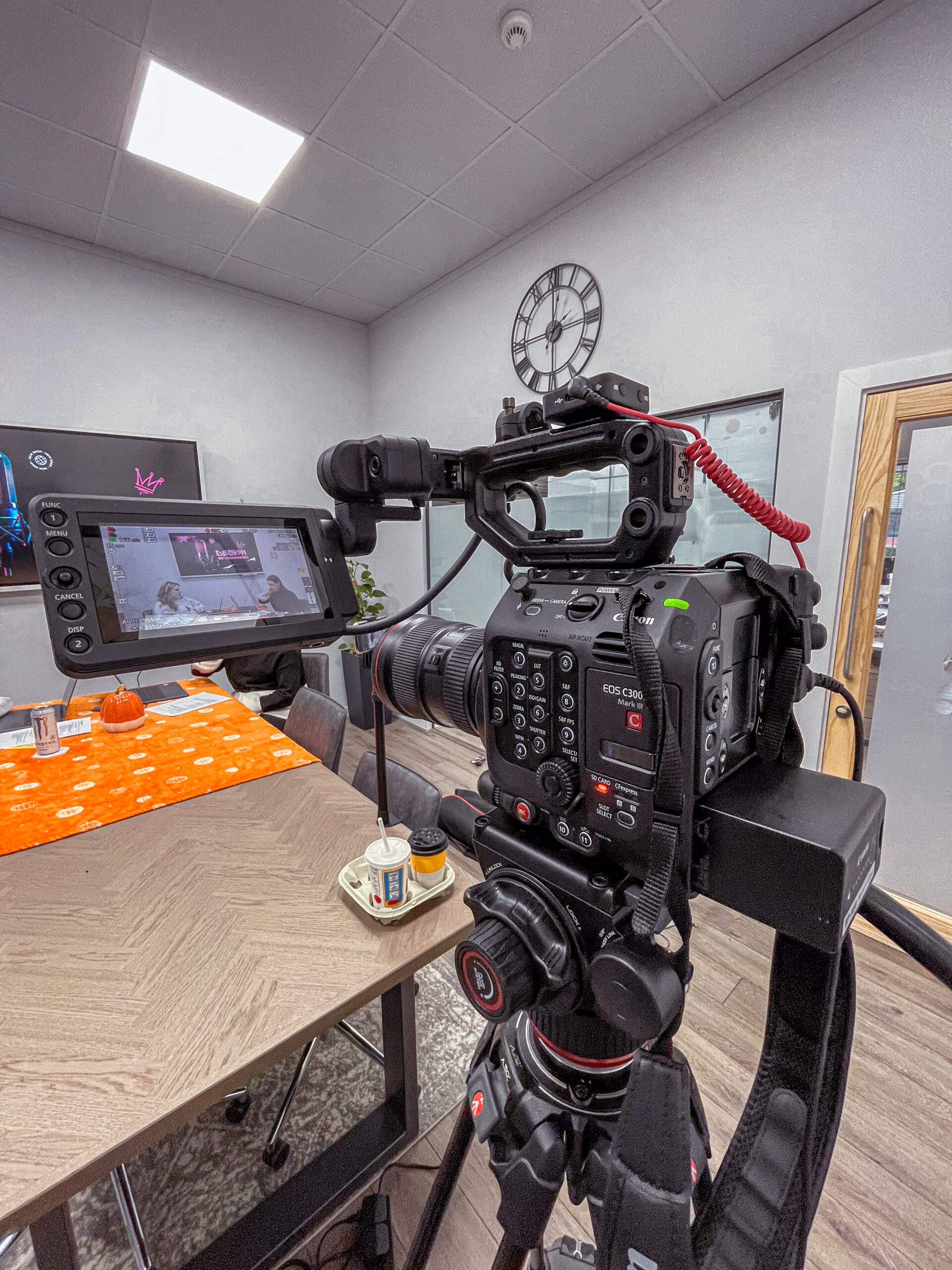 Photograph of a video camera mounted to a tripod filming Jessica Thornton and Molly Bailey while they are doing a podcast. Jessica and Molly are not directly visible but can be seen in the viewfinder eyepiece.