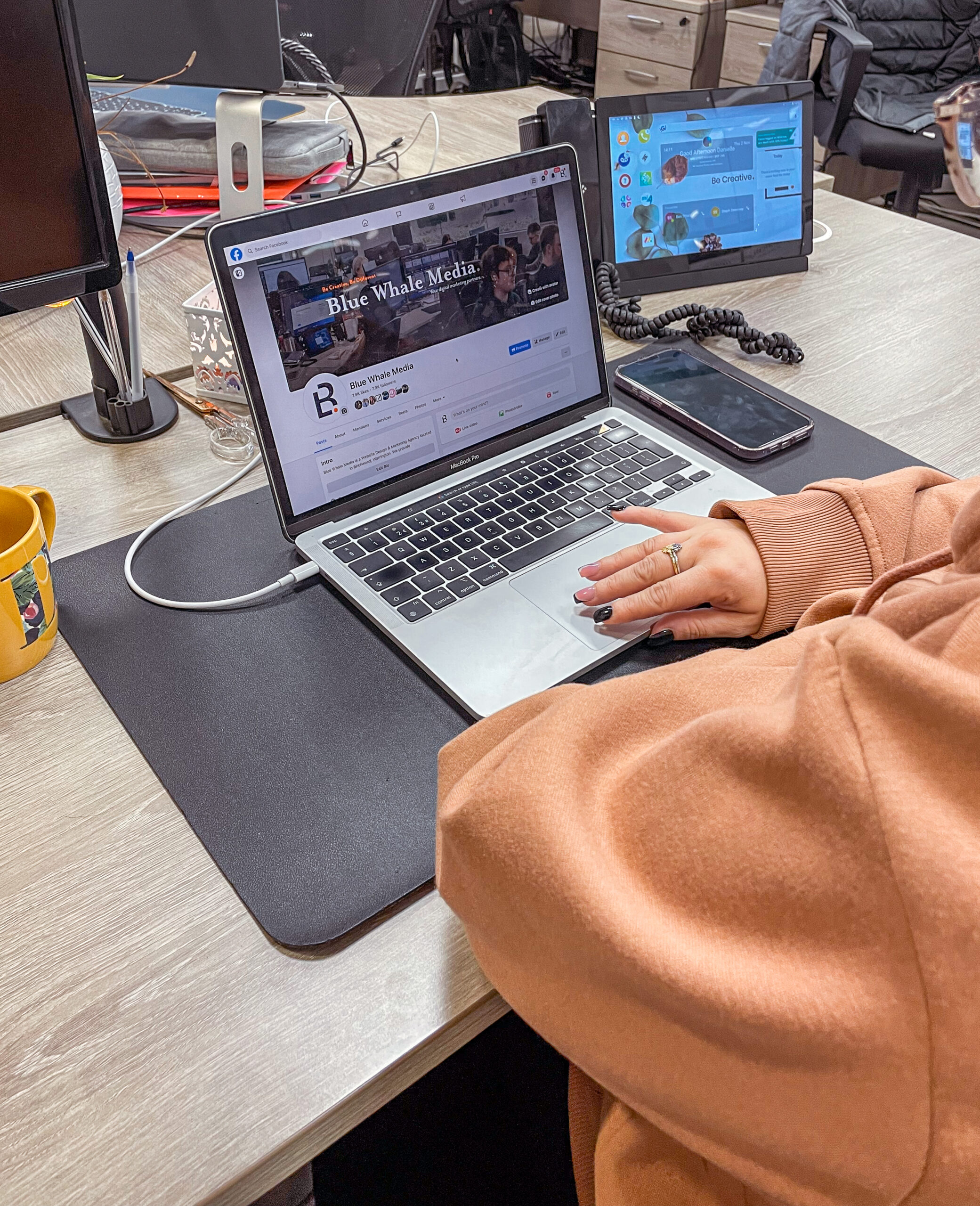 Photograph of a staff member sat at their desk working on a Macbook. On the Macbook screen is the Blue Whale Media facebook page